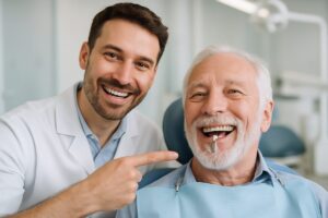 A dentist smiling and pointing to a dental implant in a patient's mouth, both are in focus and look happy. The background is blurred, clean, and modern, suggesting a state-of-the-art dental clinic. No text on the image.